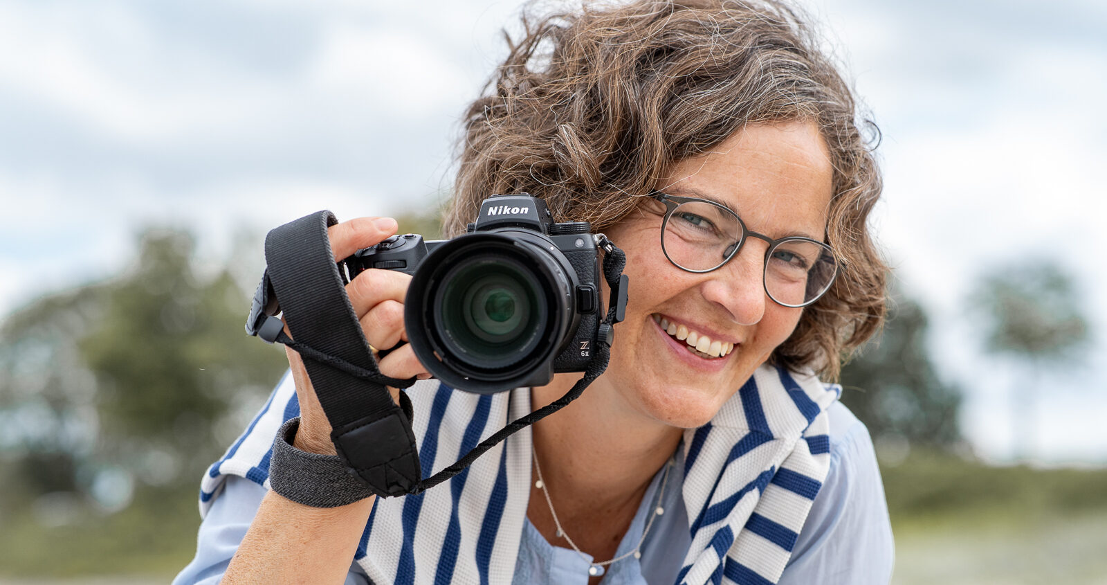 Portrait von der Fotografin Annette Kleinschmidt aus Glücksburg, Flensburg und Umgebung. Annette guckt fröhlich am Strand an ihrer hochgehaltenen Kamera vorbei.