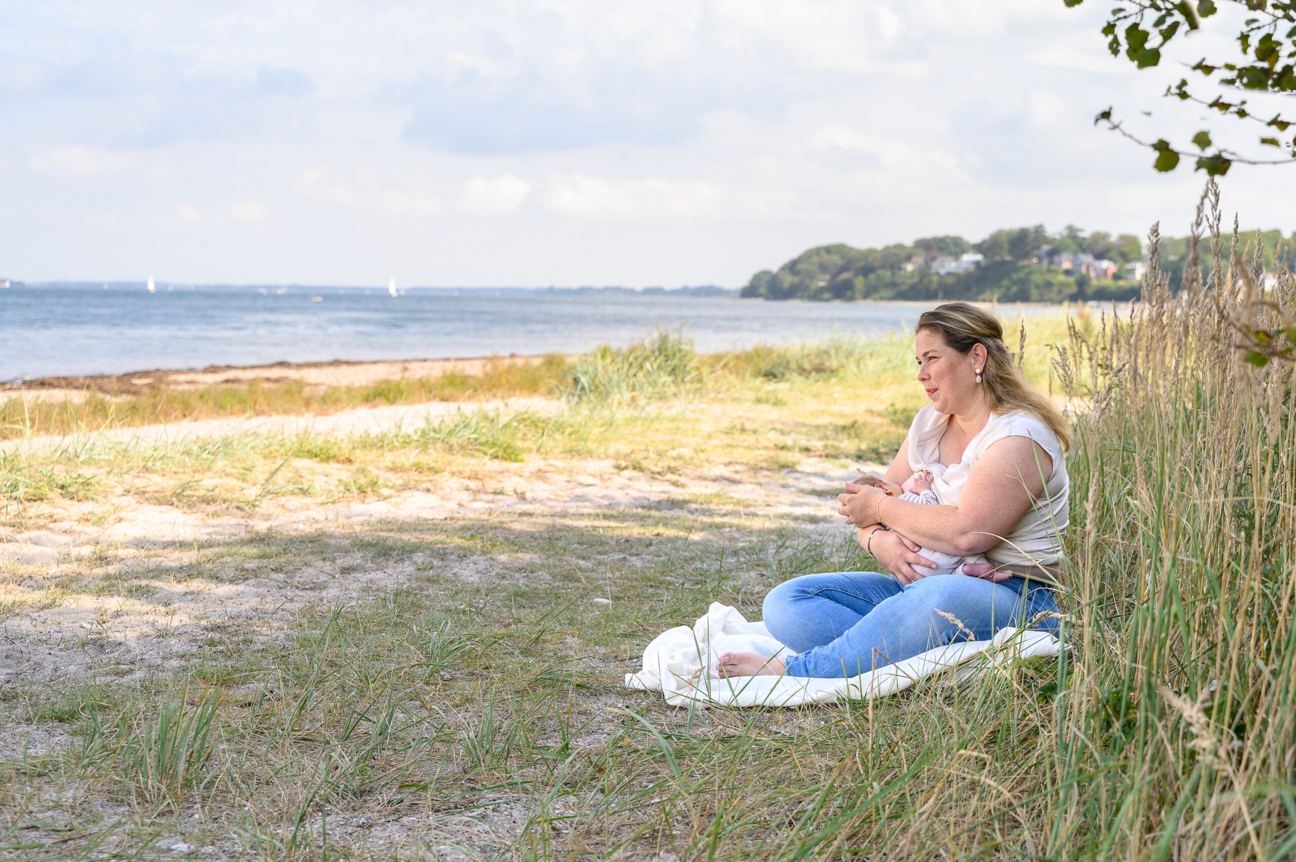 stillende Mama sitzt vertraeumt und allein am Gluecksburger Strand in Quellental und schaut auf die Flensburger Foerde in enger Verbindung mit ihrem Baby