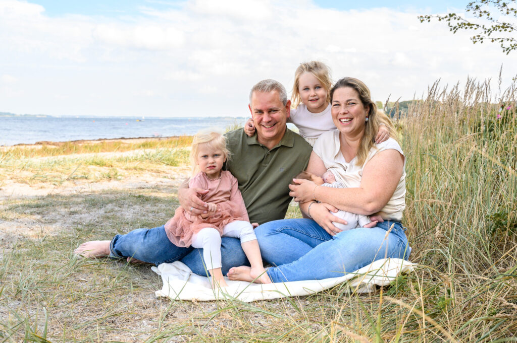 fröhliche fuenfkoepfige Familie sitzt am Strand an der Flensburger Foerde und strahlt die Fotografin an
