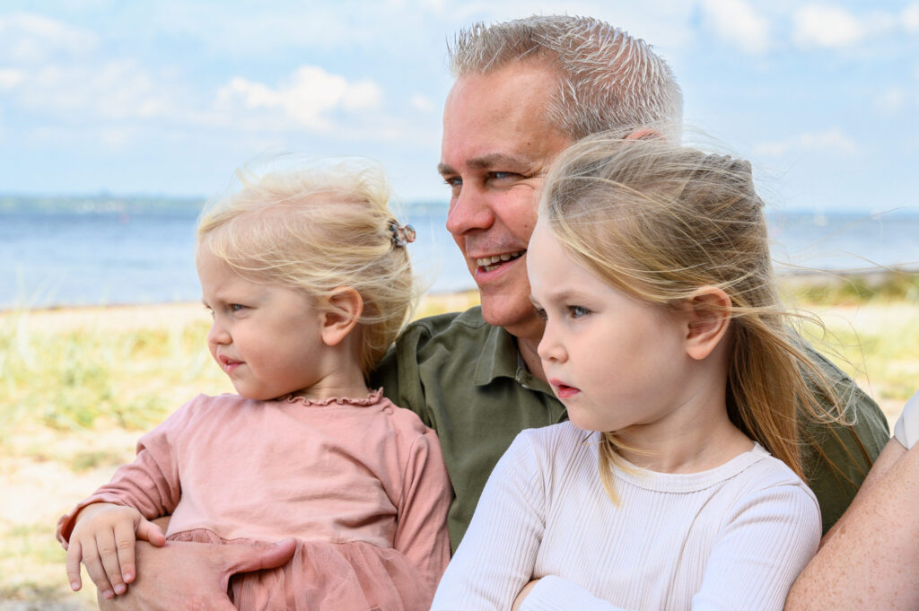 Familienfoto am Strand auf dem der Papa seine Töchter im Arm hält und alle in die gleiche Richtung auf die Flensburger Förde gucken.
