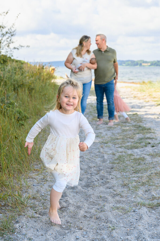 Mädchen im weissen Sommerkleid laeuft froelich auf die Fotografin zu während die Eltern im Hintergrund sich liebevoll anschauen