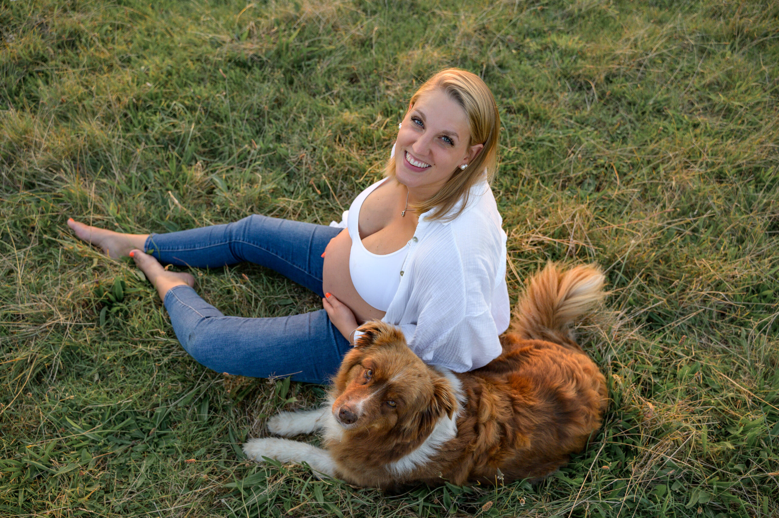 Mama sitzt im Gras und haelt ihren Babybauch und guckt laechelnd zur Fotografin hoch neben ihr liegt ihr Hund