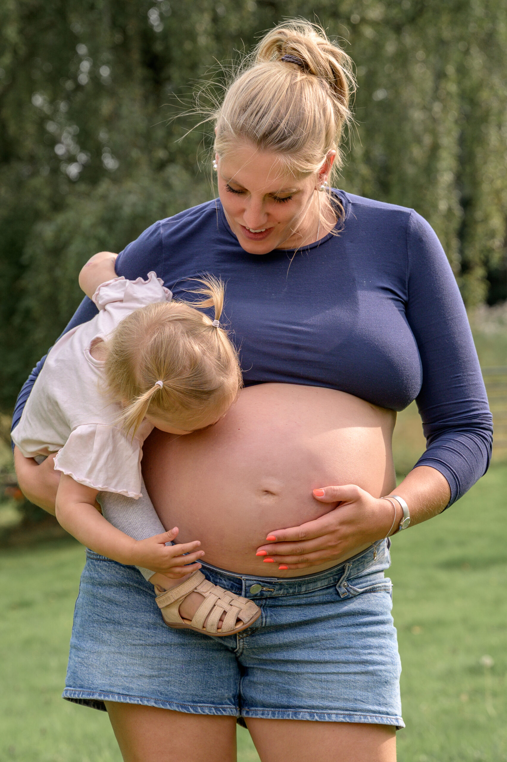 Mama in kurzer Hose hält mit einer Hand ihren Babybauch und mit der anderen ihre kleine Tochter auf dem Arm während das Mädchen den Bauch kuesst