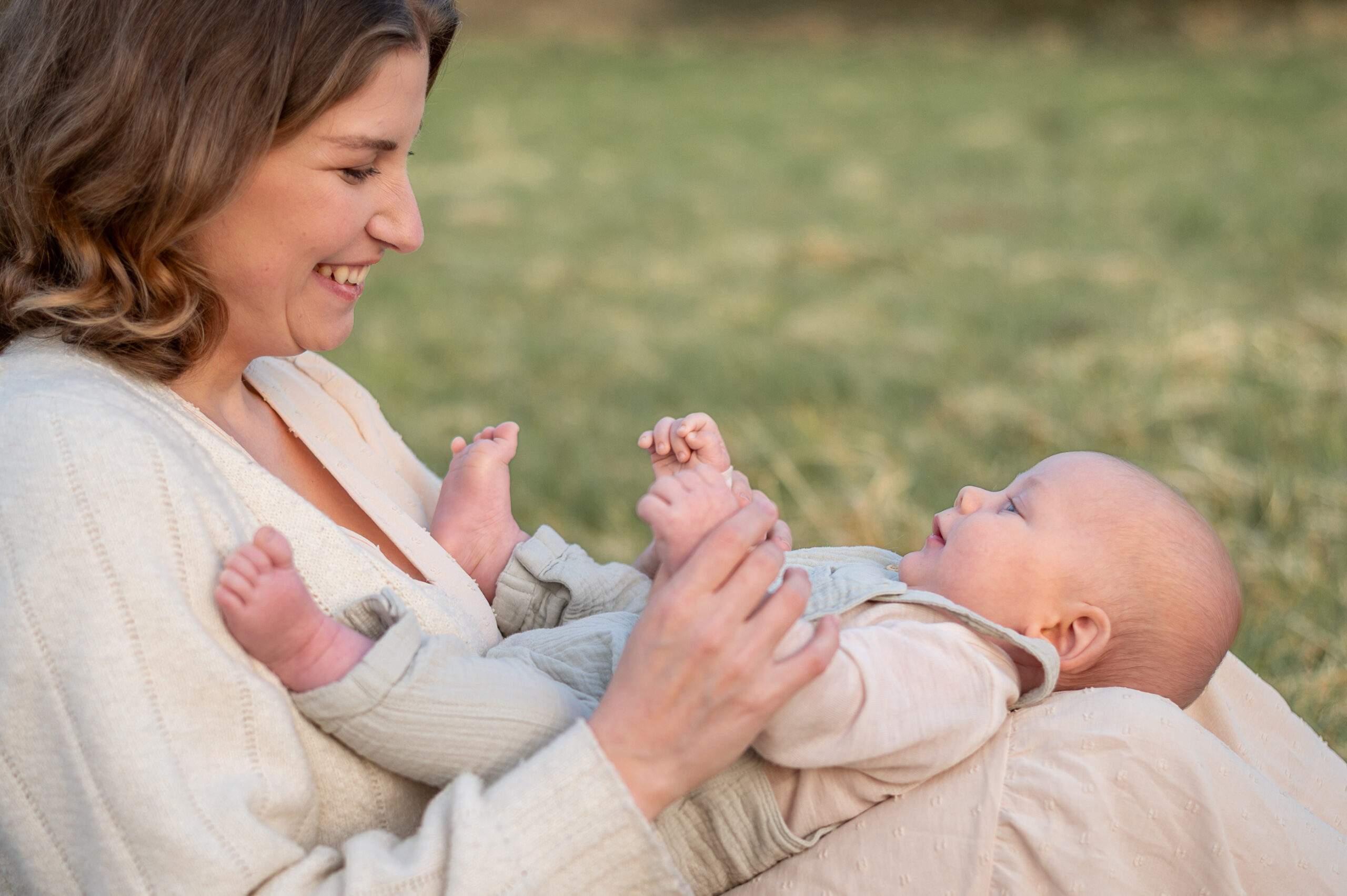 Mutter sitzt in der Natur und ihr Baby liegt auf ihren Beinen beide schauen sich laechelnd anund halten sich bei der Hand