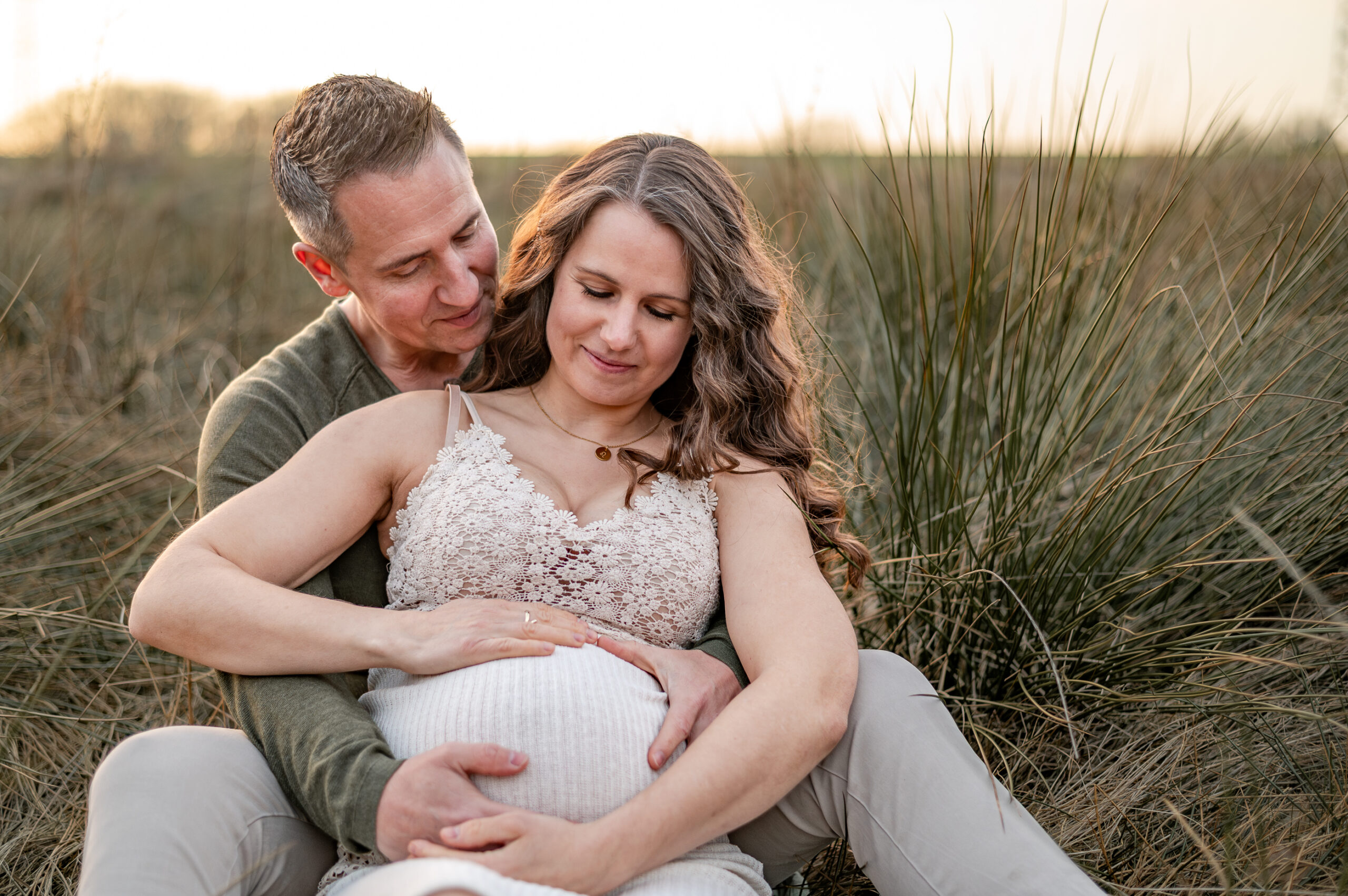 Schwangere lehnt sich im Sitzen in der Natur in Flensburg an ihren Partner, beide halten den kugeligen Bauch und schauen voller Vorfreude auf den Bauch