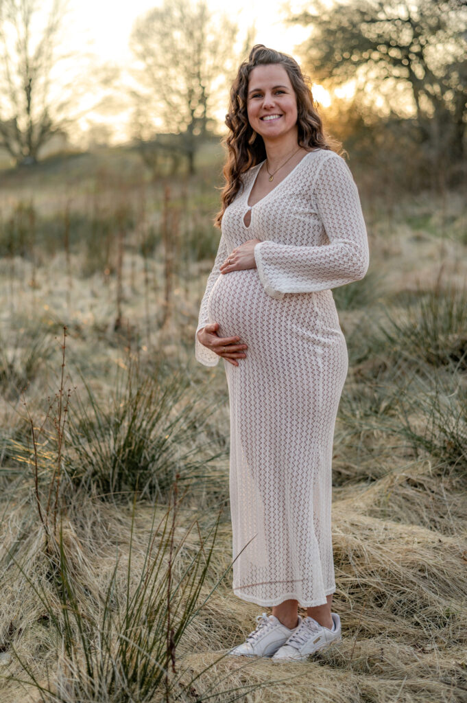 Schwangere im weissen langem Kleid schaut gluecklich laechelnd in die Kamera und haelt ihren Babybauch