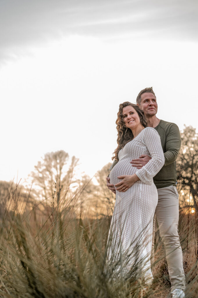 Das Paar schaut gemeinsam in die Weite der Natur und strahlen dabei Vorfreude auf den Familienzuwachs aus. Das Babybauchfoto ist aus der Froschperspektive aufgenommen und laesst den Bauch natürlich und kugelig wirken.