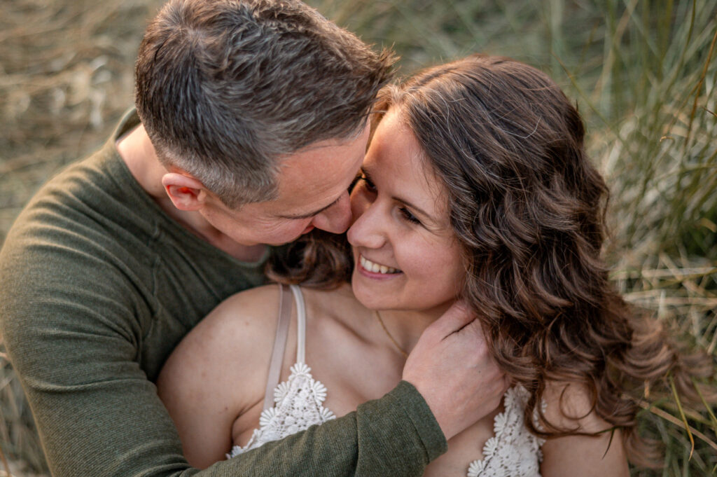 Paar- und Portraitfoto beim Babybauchshooting in der Natur auf einer Wiese in Flensburg. Die werdenden Eltern halten sich eng im Arm und schauen sich innig und nah in die Augen.