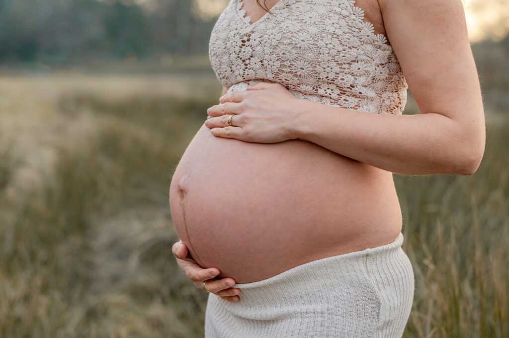 ein unbekleideter Babybauch von den Haenden der werdenden Mutter von oben und unten gehalten. Die Mama steht auf einer Wiese in der Natur.