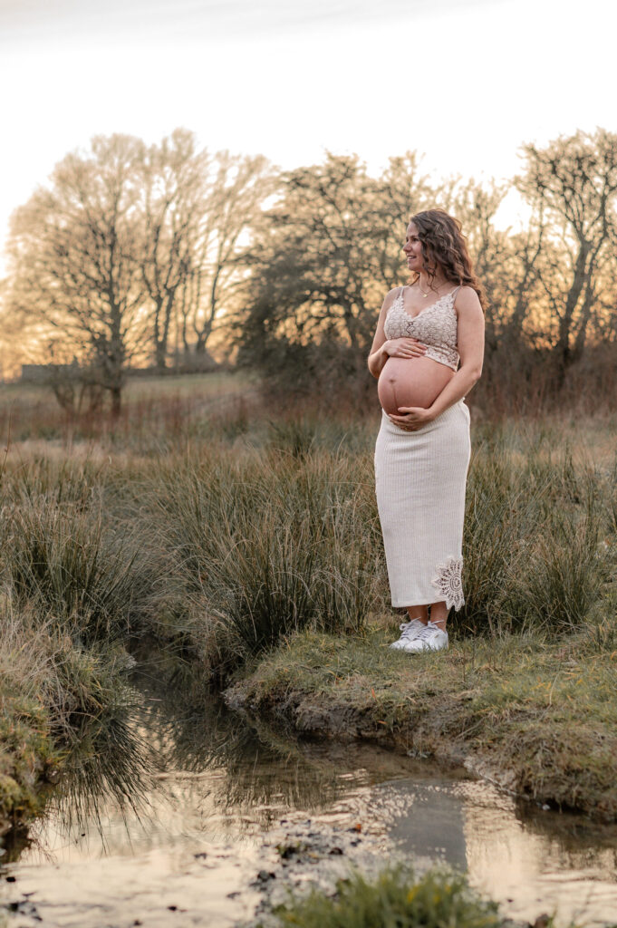 naturliches Babybauch Shooting an einem kleinen Bach steht die werdende Mam und haelt ihren Bauch.