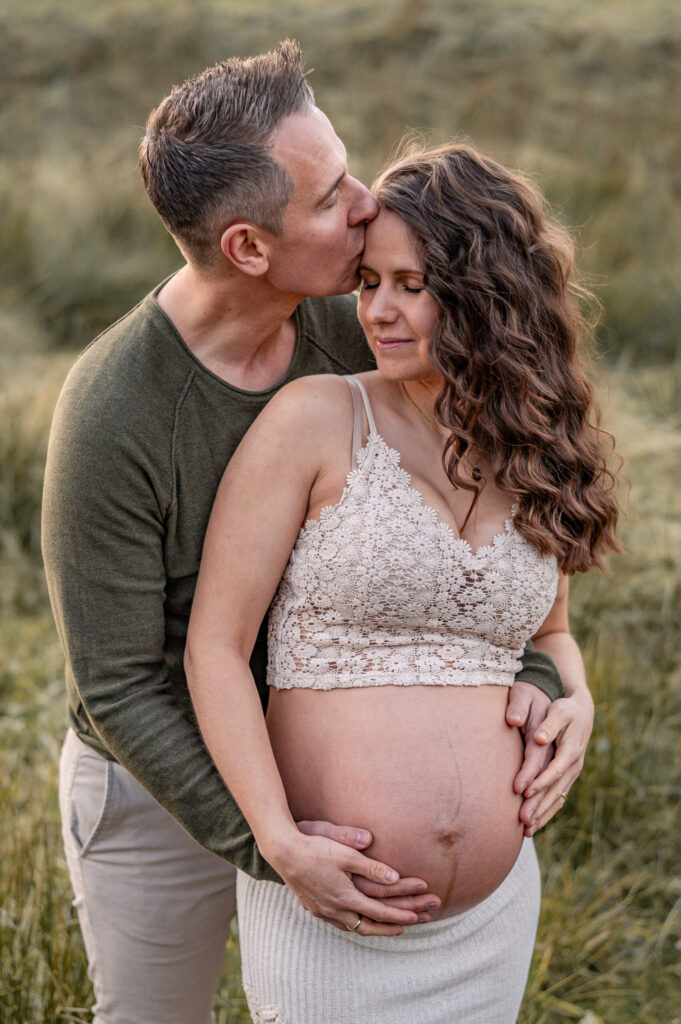 Werdende Eltern beim Babybauch Shooting in der Natur in Flensburg. Der werdende Vater umarmt seine Partnerin und den Schwangeren Bauch von hinten und küsst ihre Stirn. Das Foto zeigt eine enge Verbundenheit und Vorfreude auf das neue Leben.