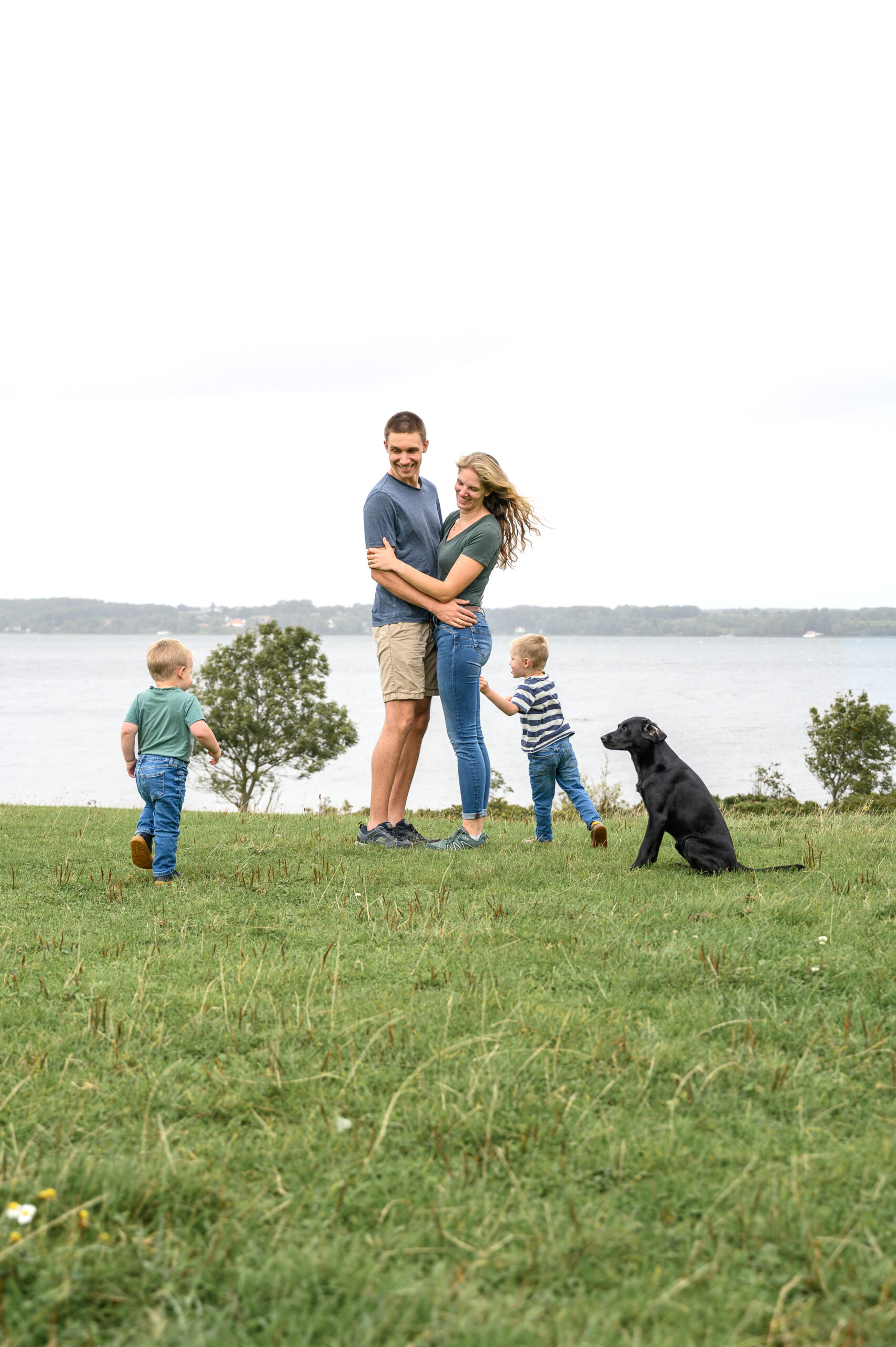 Familien Fotoshooting auf einer Wiese mit Blick auf die Flensburger Foerde. Die beiden kleinen Soehne und der schwarze Hund laufen froehlich um die Eltern herum, die sich umarmen.