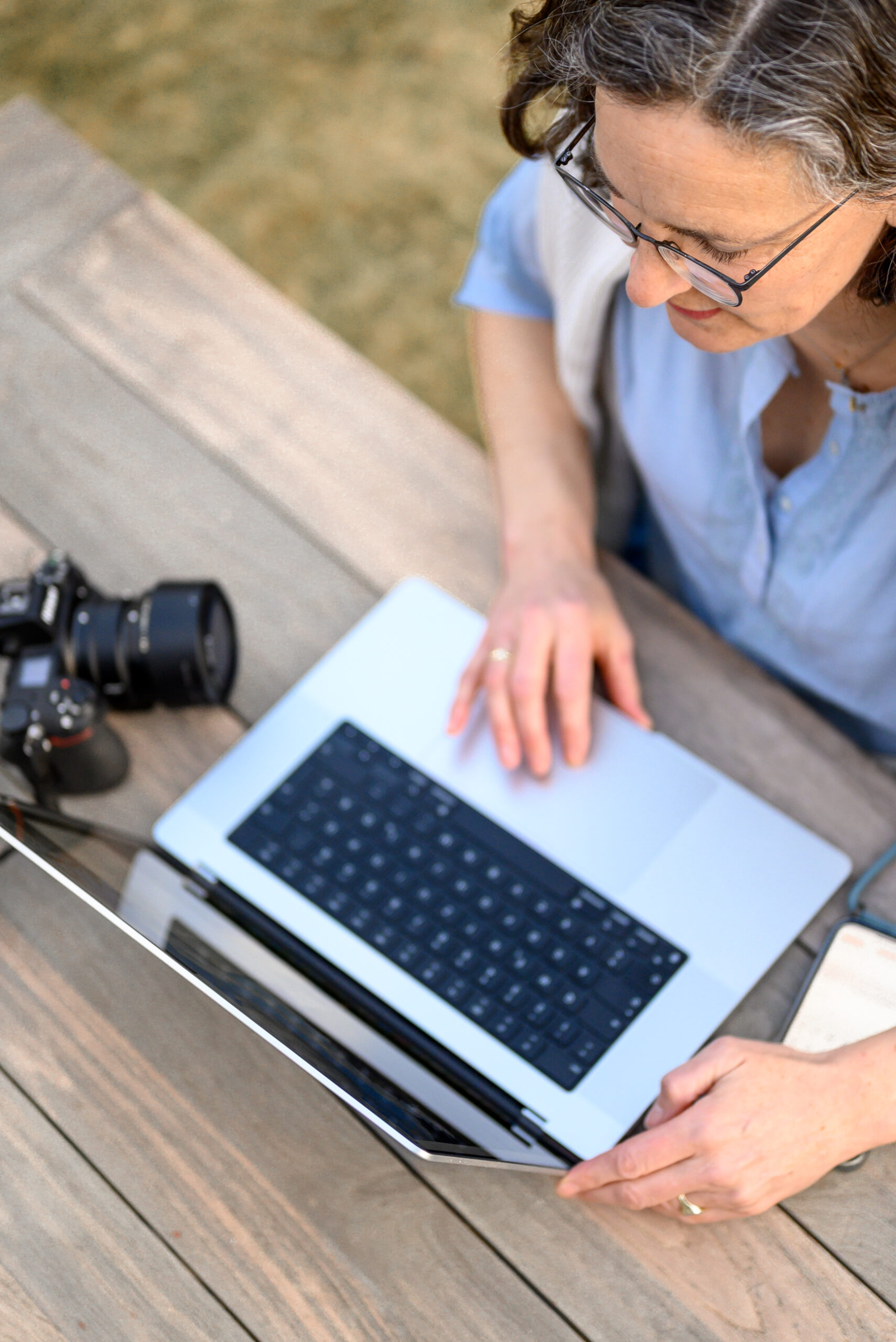 Annette Kleinschmidt von Fotografie Gluecksburg sitzt arbeitend an ihrem Computer draussen im Garten an einem Tisch. Neben dem Computer ist auch noch die Fotokamera und das Mobiltelefon zu sehen.