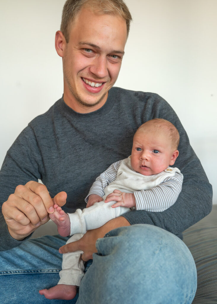 Papa sitzt mit Baby auf dem Schoss beim Baby Shooting als Homestory in Gluecksburg. Der Papa schaut entspannt in dir Kamera und sein Baby Sohn liegt gemütlich mit offenen Augen in Papas Arm.