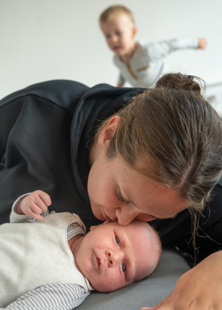 Mama im Wochenbett gibt Neugeborenem einen zärtlichen Kuss auf den Kopf. Bruder spielt im Hintergrund.