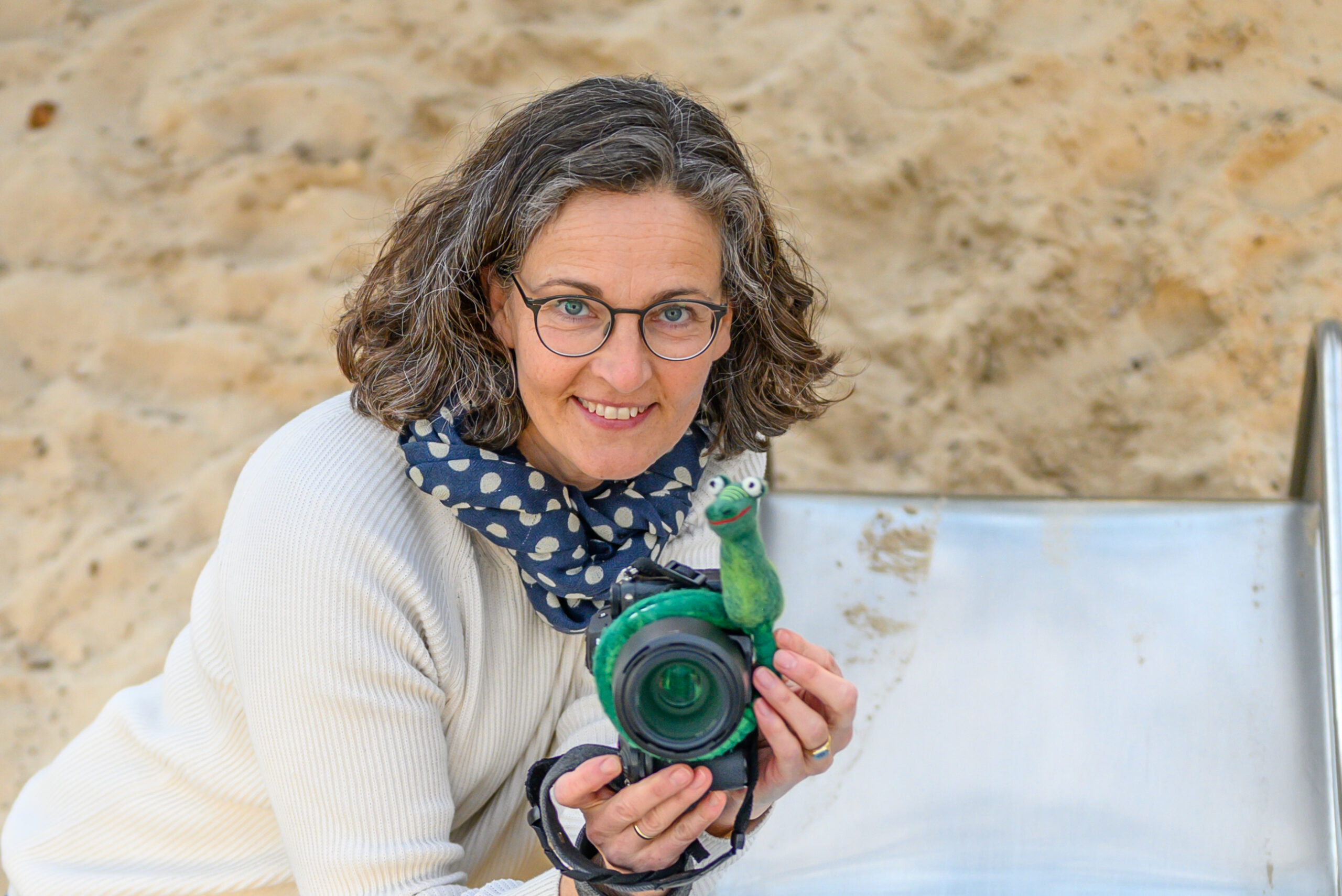 Annette Kleinschmidt hockt als Kita-Fotografin in Gluecksburg, Flensburg und Umgebung mit ihrer Kamera am Ende der Rutsche draußen auf einem Spielplatz und fotografiert Kitakinder. Annette lächelt entspannt in die Kamera im Hintergrund sieht man das Ende Rutsche und viel Sand.