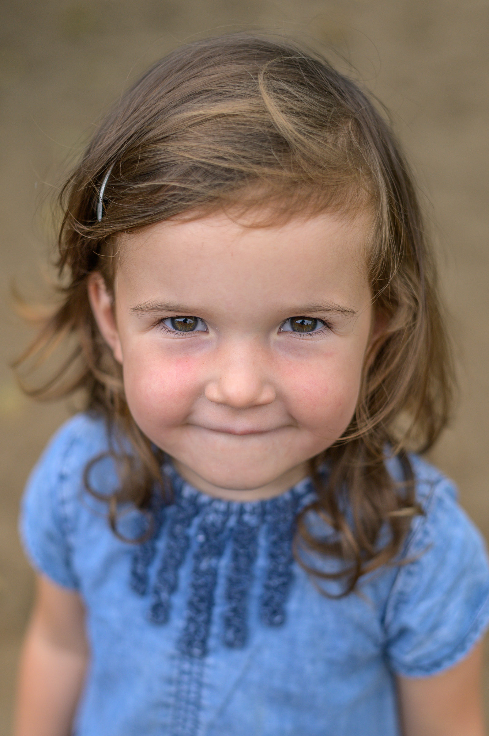 Kindergartenkind im blauen Kleid schaut am Fototag von unten direkt in die Kamera. Die Fotografin Annette Kleinschmidt aus Glücksburg, Flensburg und Umgebung spiegelt sich in den Augen des Mädchens.