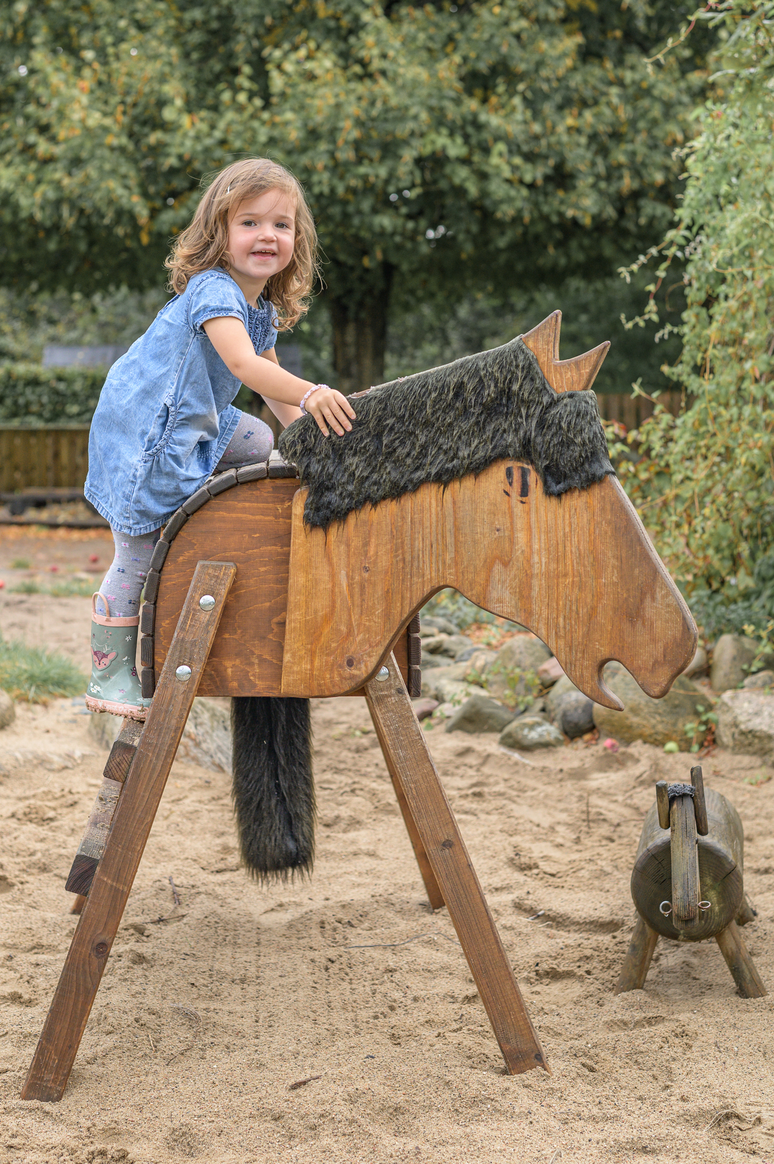 Mädchen klettert in Gummistiefel und Kleid auf ein Holzpferd im Aussenbereich der Kita in Flensburg und Umgebung.