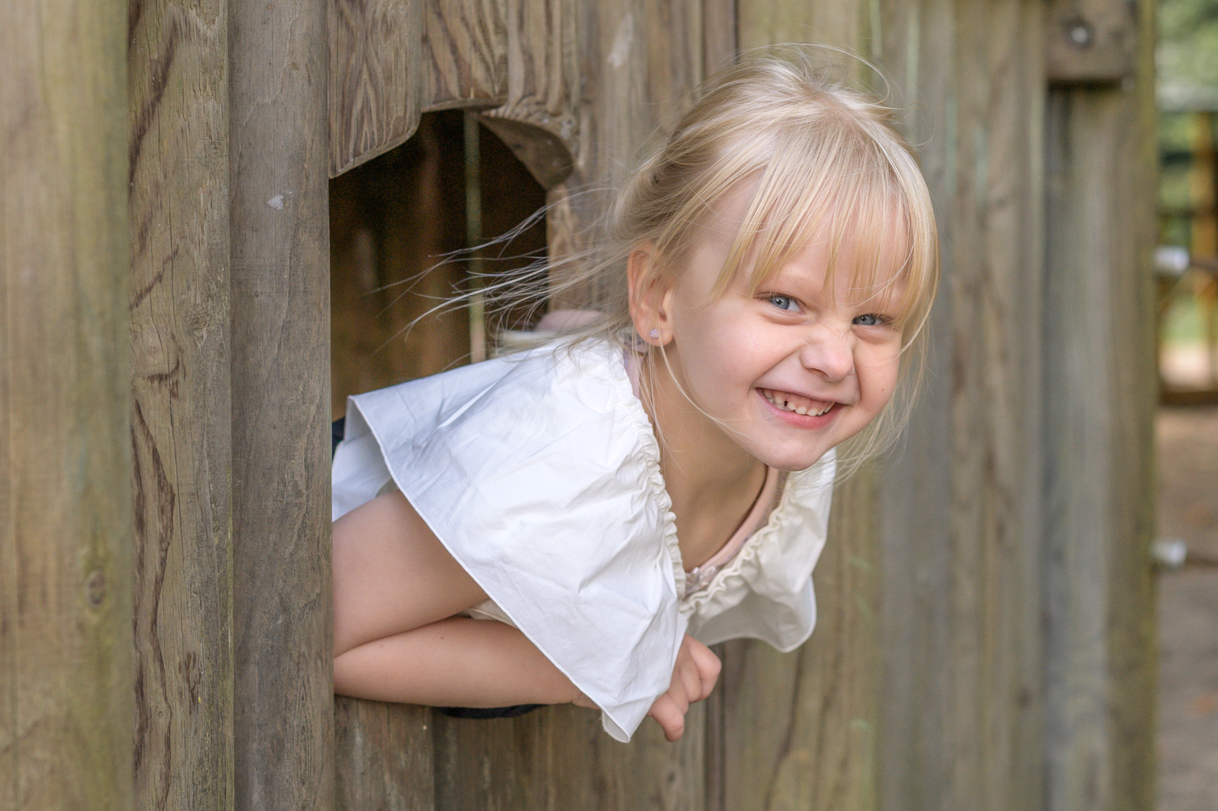 frech guckendes Kita Mädchen guckt aus dem Fenster des Holzhauses auf dem Kindergarten Spielplatz und grinst in die Kamera.