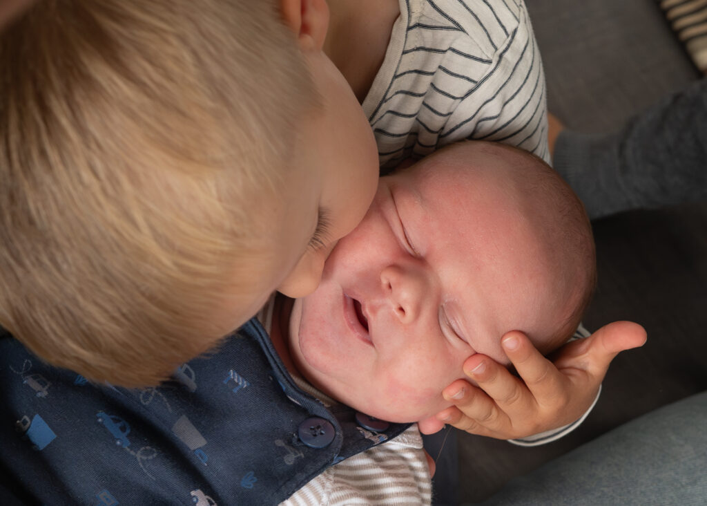 während des Baby Shooting mit Annette Kleinschmidt aus Glücksburg gibt das Geschwisterkind seinem neugeborenen Bruder einen Kuss und hält das Köpfchen vorsichtig. Das Bild ist von oben fotografiert.