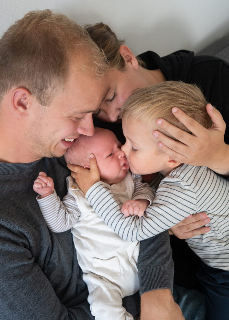 Familie mit Neugeborenen und Kleinkind kuschelt eng auf dem Sofa bei sich zu Hause in Gluecksburg. Der kleine grosse Bruder kuesst das Baby zaertlich. Das Foto zeigt ganz viel Naehe, Liebe und Verbundenheit.